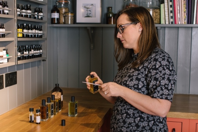 woman standing a work benches with perfume bottle and ingredients