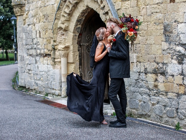 bride and groom dressed in black kissing outside of church