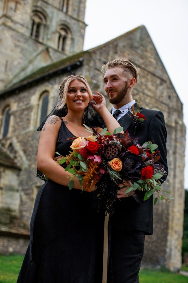 bride and groom dressed in black, outside church, bride holds beautiful red and peach bouquet