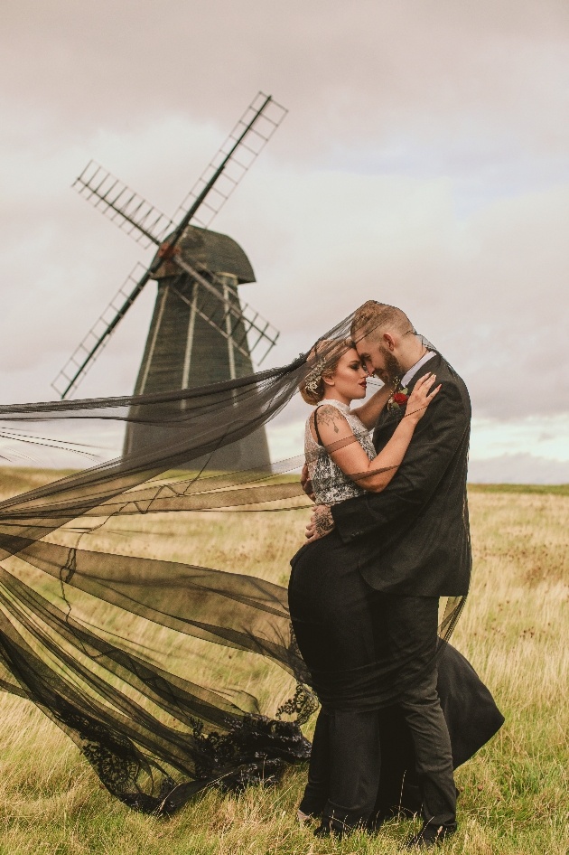 Bride and groom in black in front of windmill. Wind has caught bride's veil which flows out behind her