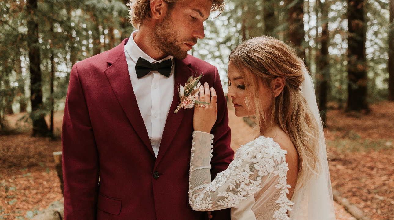 Bride and groom groom wears burgundy shoot and black bowtie both are surrounded by woodland