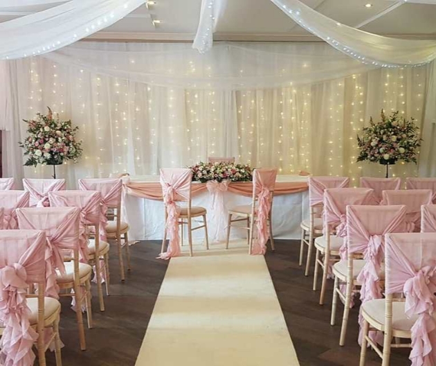 Wedding ceremony area with fairylight backdrop, pink palette, floral pedestral arrangements an blush chair covers.