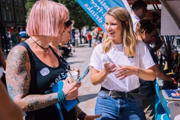 two women stand in road chatting with drinks surrounded by fair stalls