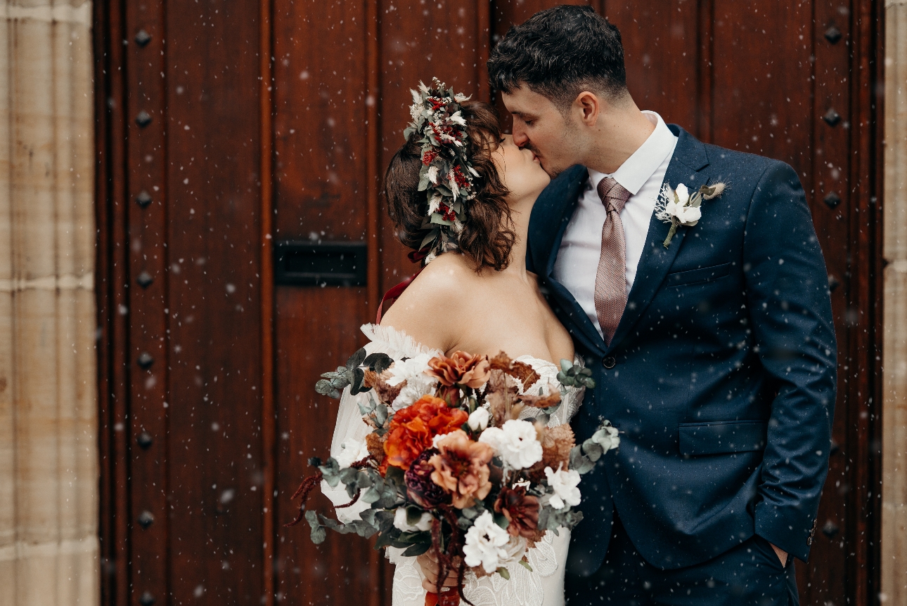 Bride and groom kissing in front of backdrop of Tonbridge School main wooden door as snow falls