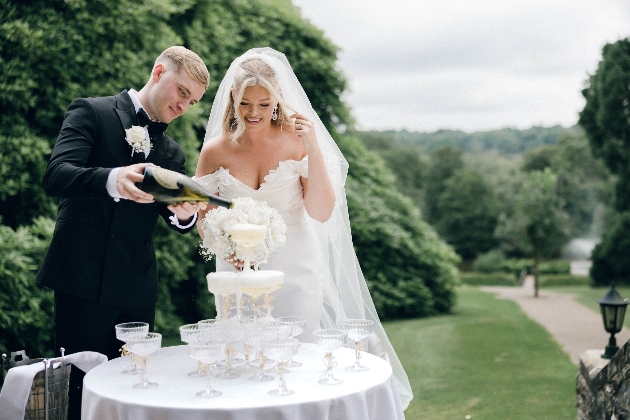 Bride and groom pouring champagne into a champagne tower with lake at Ashdown Park in the background