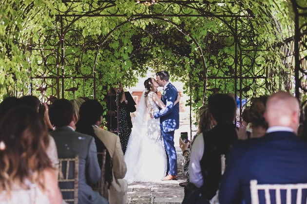 bride and groom, wedding ceremony, kissing in pergola as guests look on